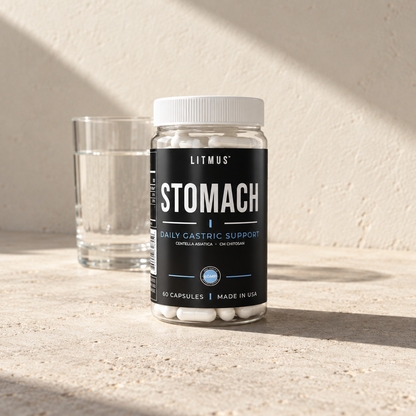 LITMUS STOMACH jar, 60 capsules, beside a glass of water on pale stone against a sunlit plaster wall.